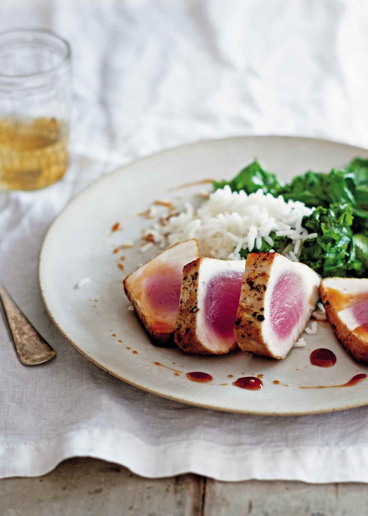A white plate with four slices of seared tuna with sweet and sour sauce, rice, and cooked greens.