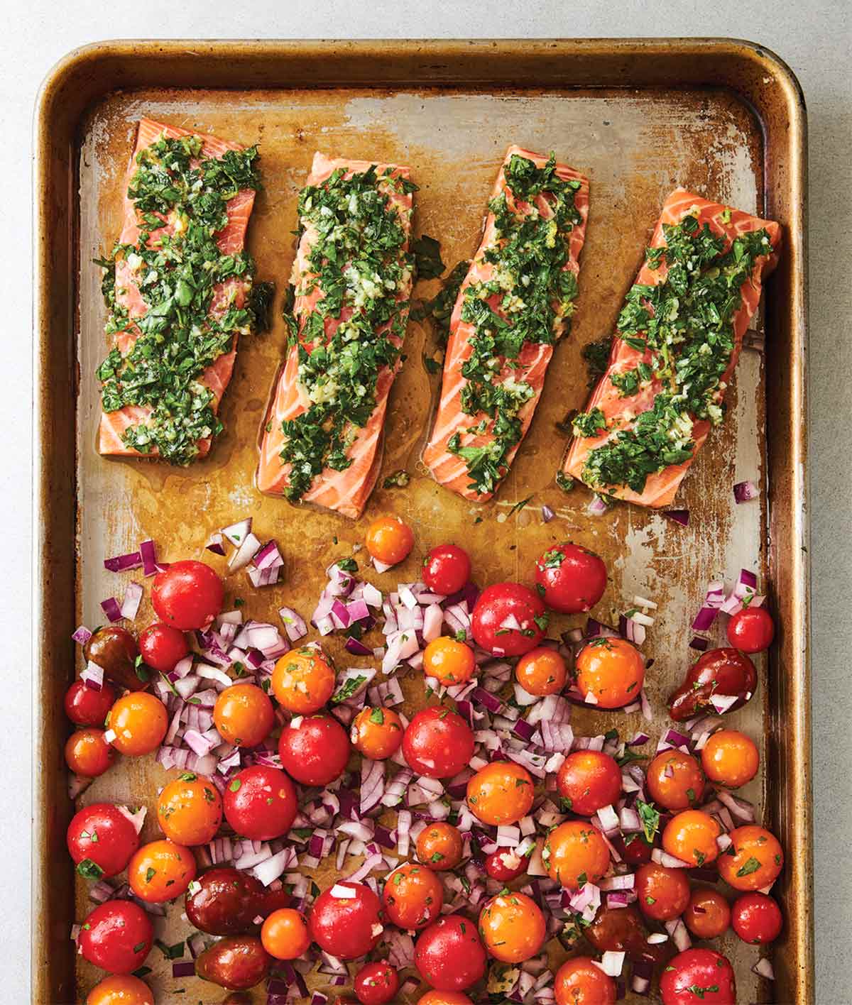 A metal sheet pan with 4 salmon fillets covered with gremolata, beside a pile of cherry tomatoes and diced red onions.