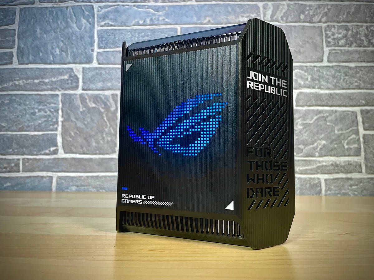 A single Asus ROG Rapture GT6 mesh gaming router sits in front of a stonework background, with the LED array displaying the ROG logo in blue.