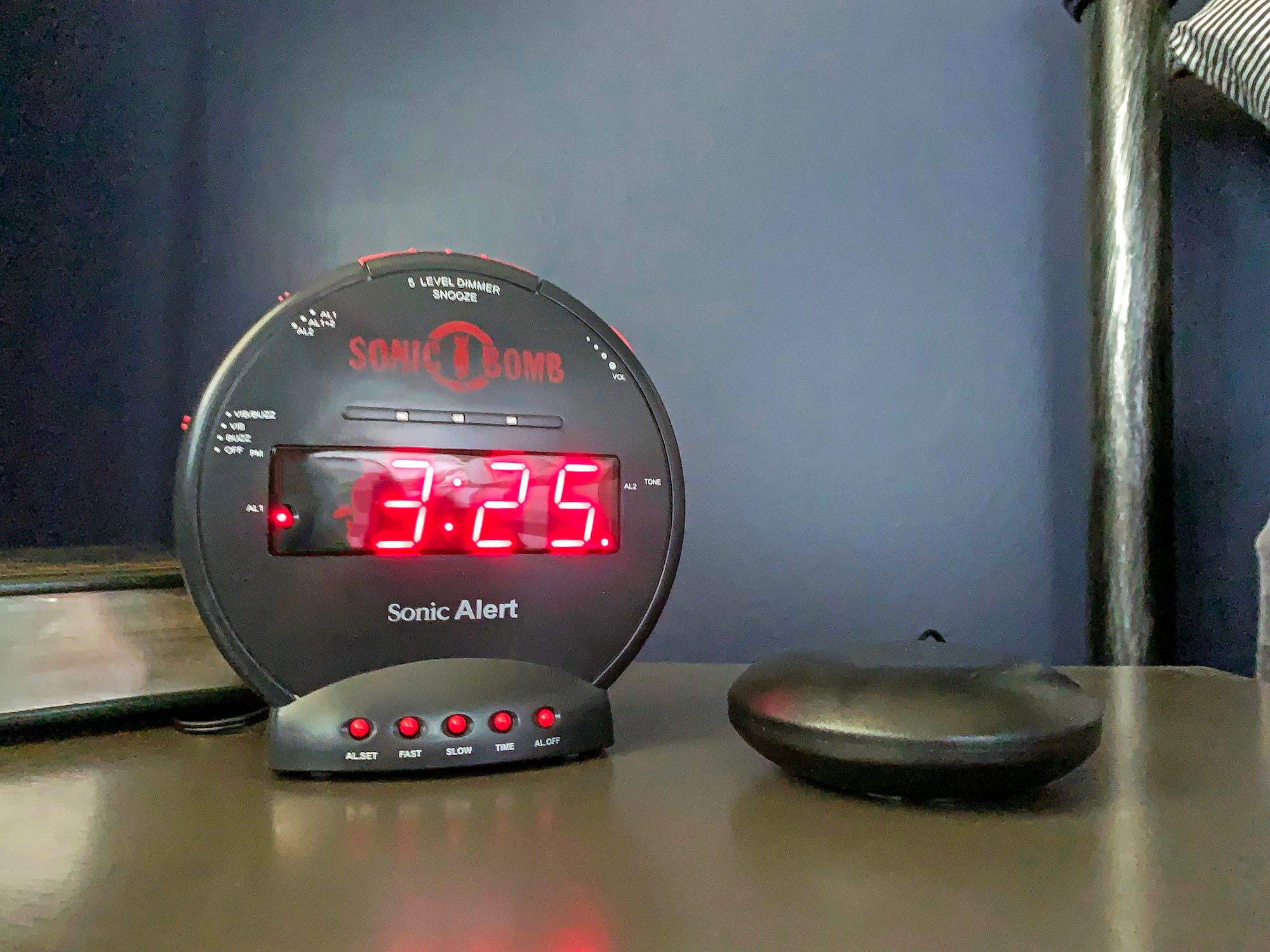The Sonic Alert Sonic Bomb Dual Extra-Loud Alarm Clock with Bed Shaker sits on a nightstand and reads 3:25.