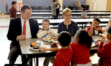 man and a woman sitting at a table in a cafeteria with school children in red shirts