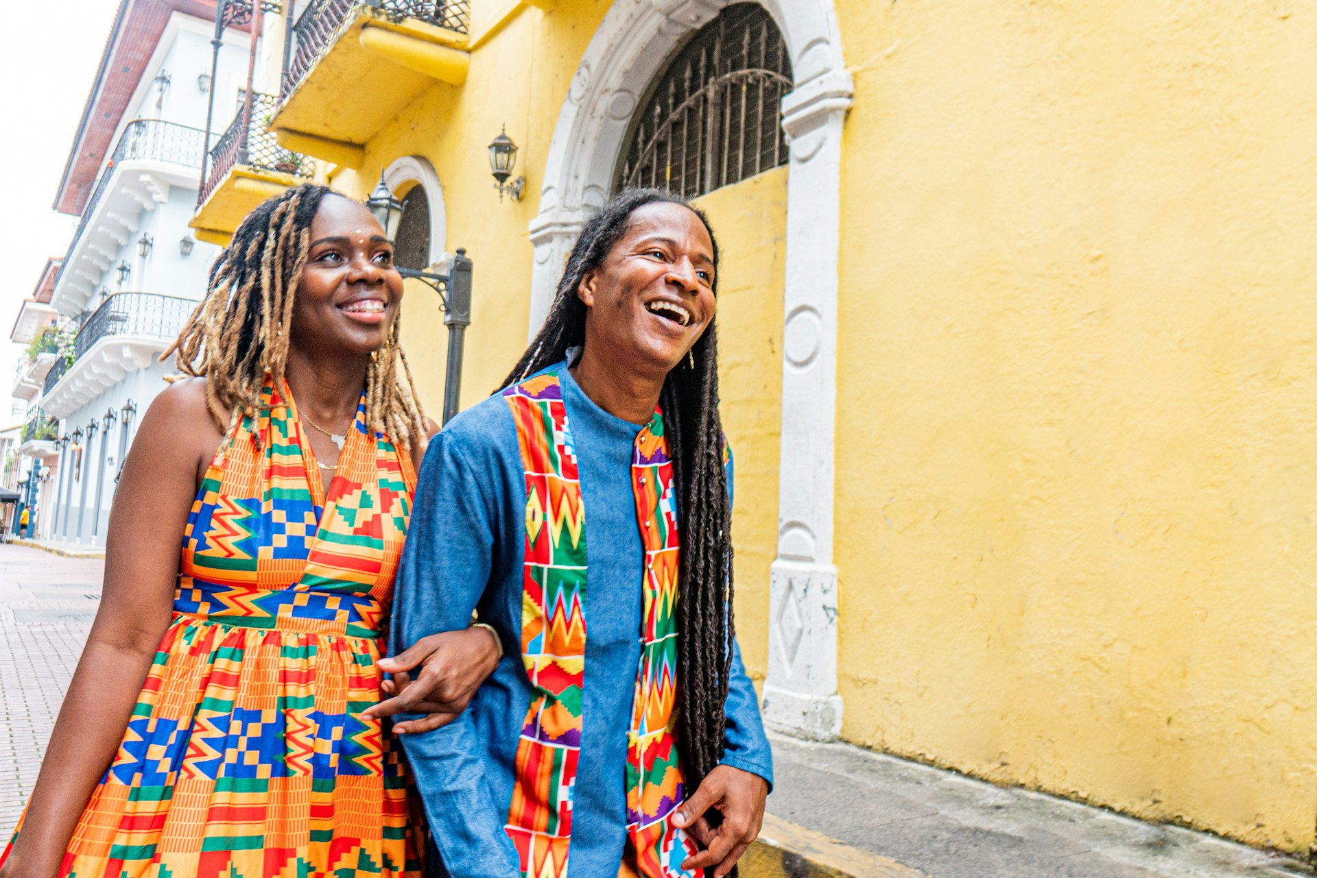 Two people wearing brightly colored clothing walk down a street smiling