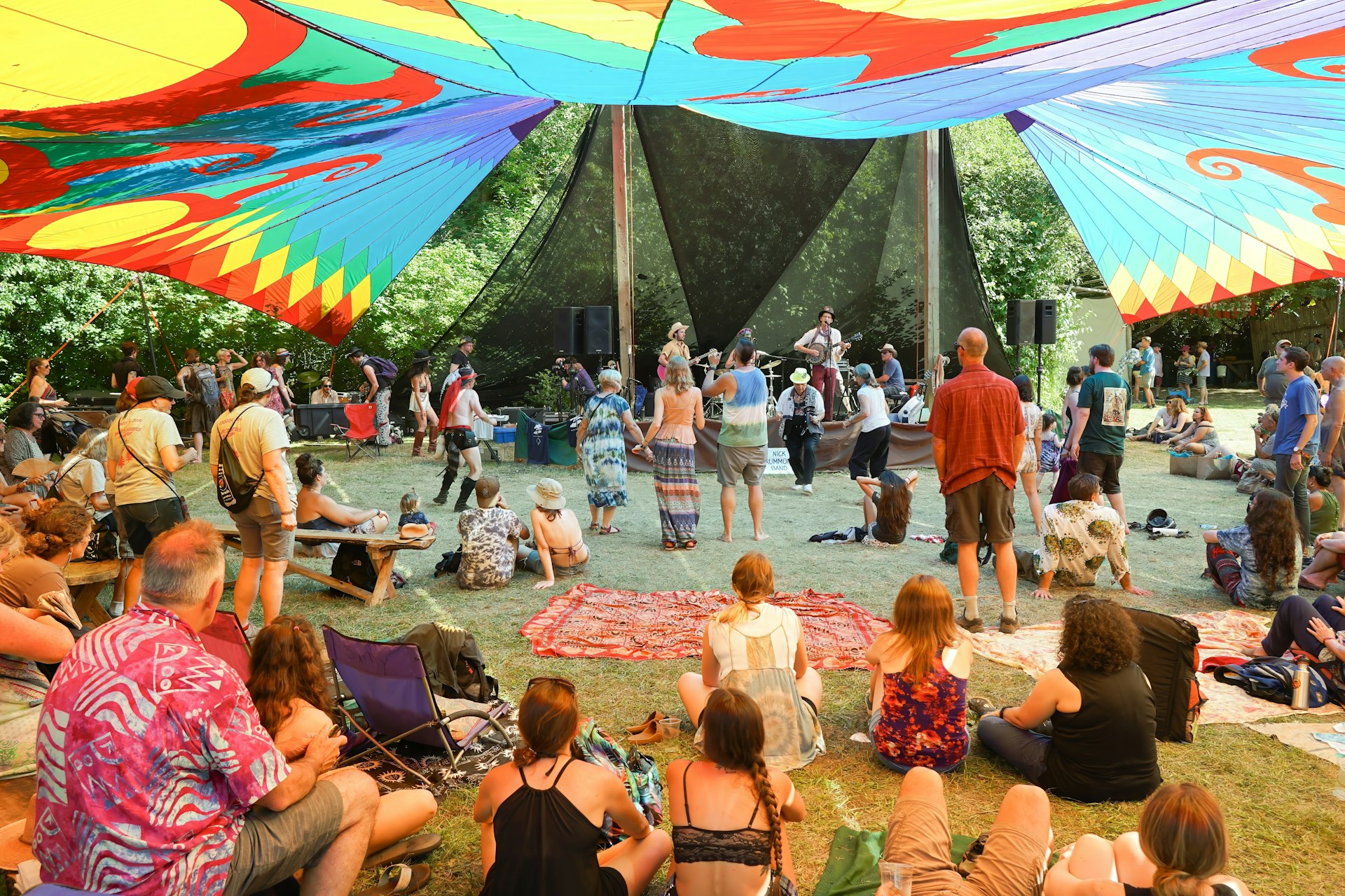 Adults and children sit under a colorful tie-dyed canopy watching a musician perform on a sunny day