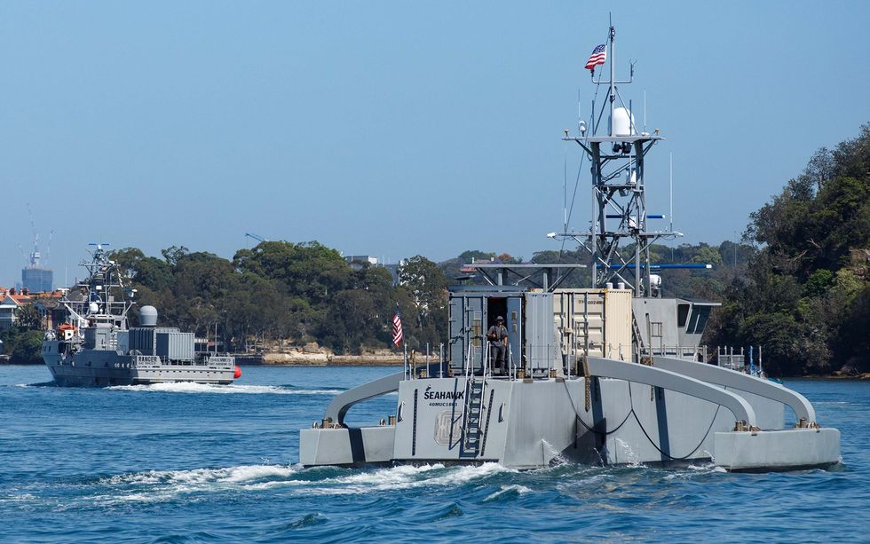 A battleship-gray trimaran ship cruises near a wooded shoreline.