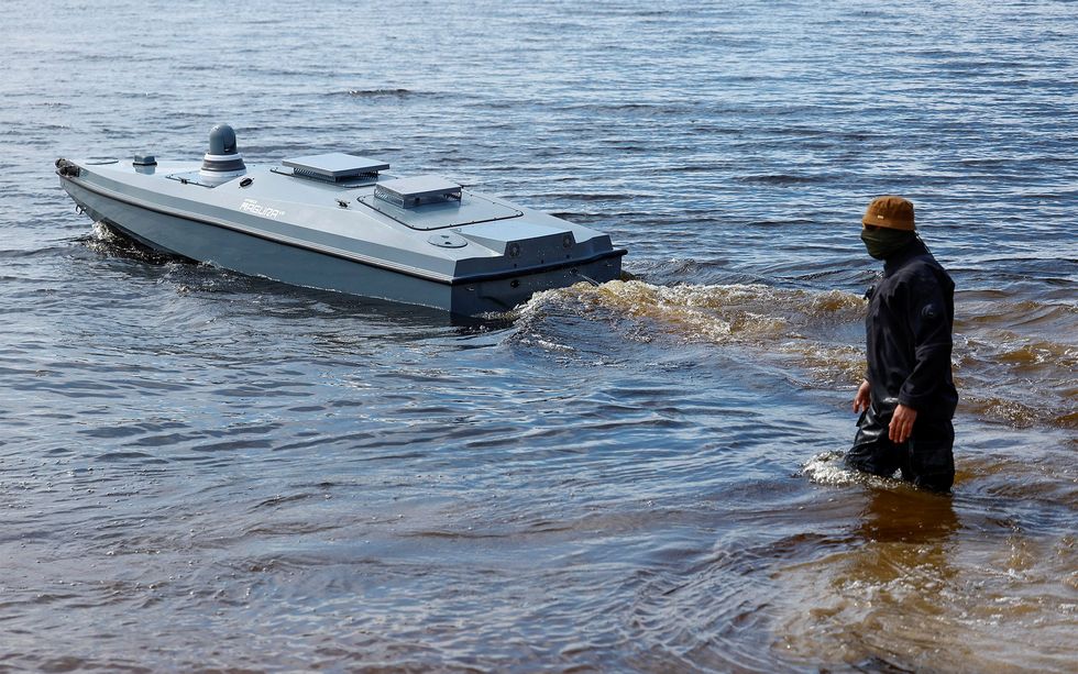 A man in a black wetsuit and brown bucket hat stands in shallow water next to a gray naval drone. 