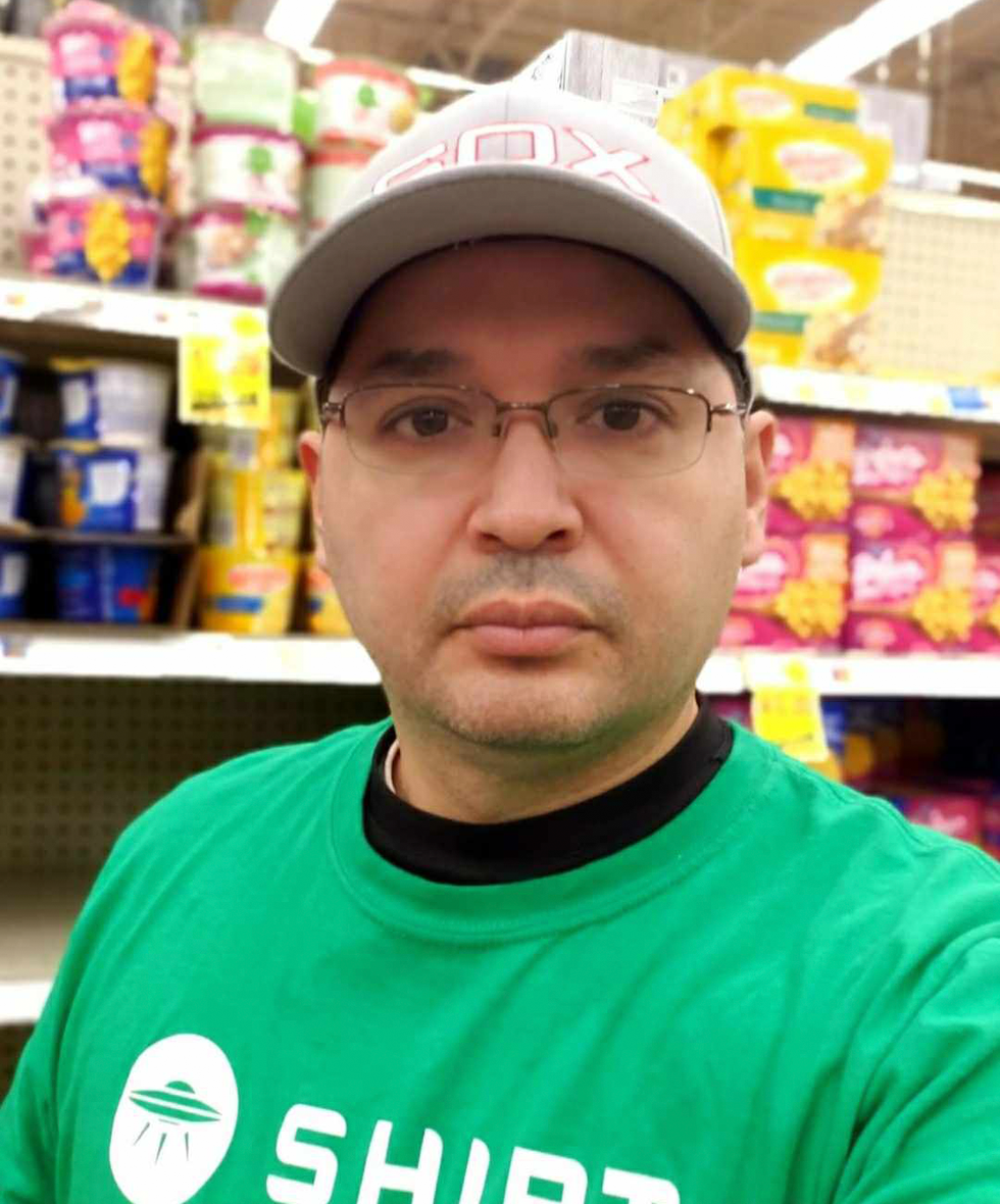 A man in a green shirt and white baseball cap looks into the camera. Heu2019s in the aisle of a grocery store.