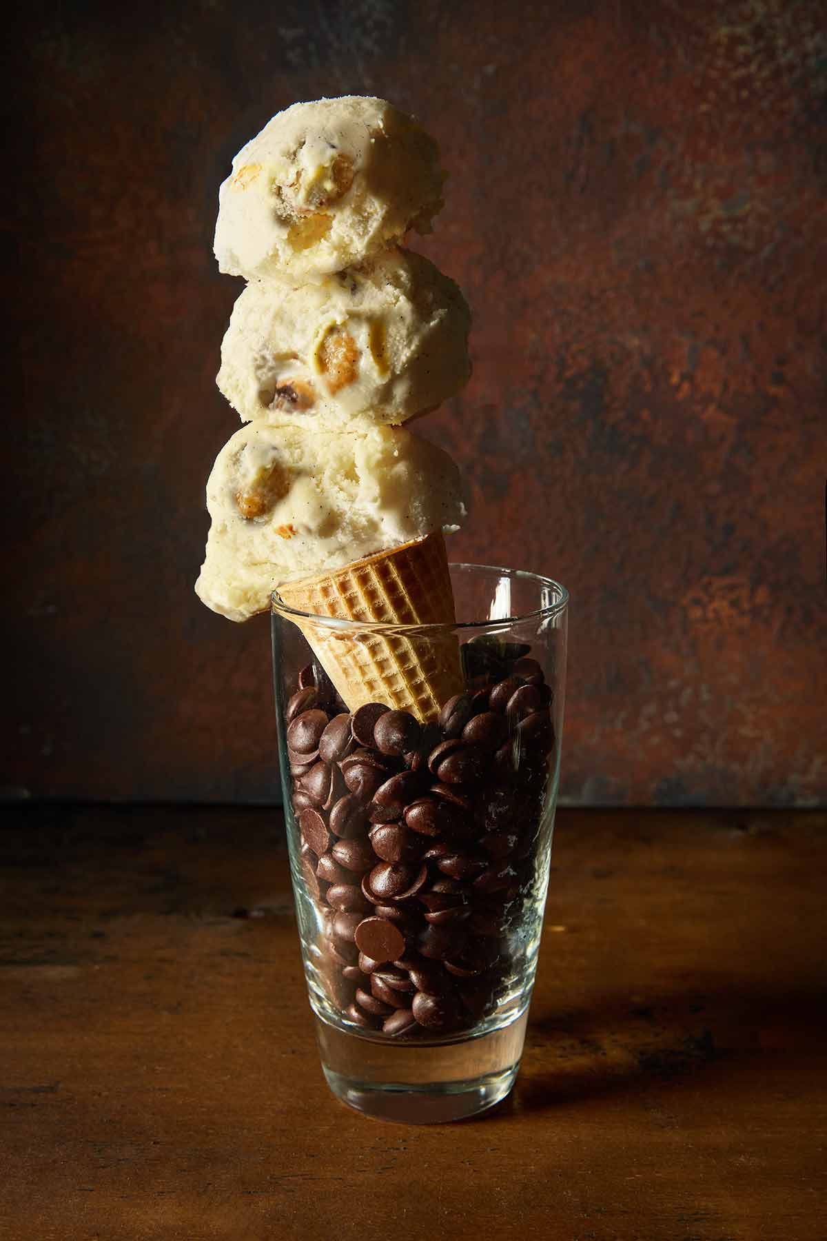 A sugar cone of chocolate chip cookie dough ice cream in a tall glass of chocolate chips.