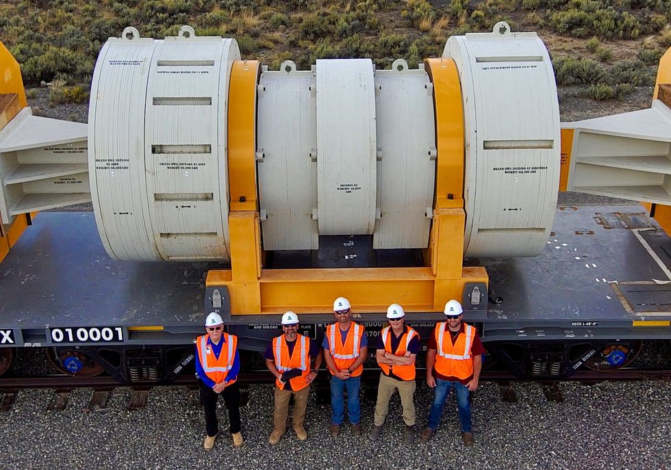 Five workers in vests and hard hats stand in front of a flat rail car. Behind them looms a larger horizontal container.