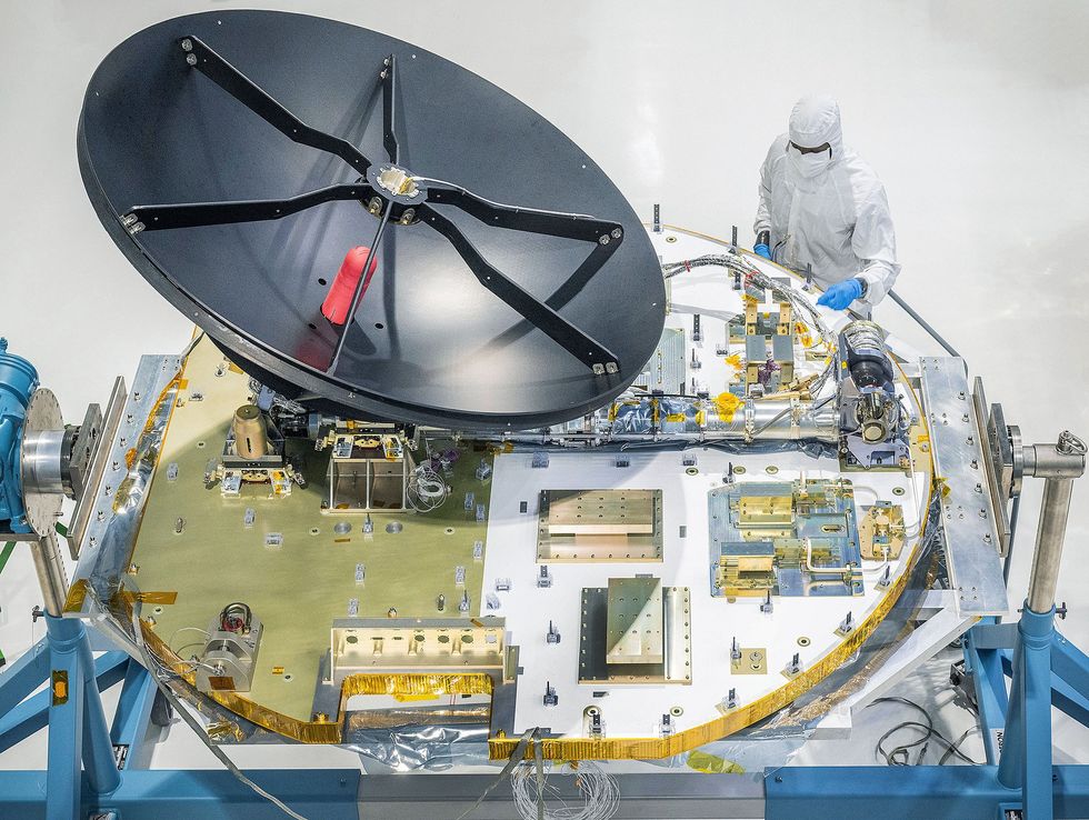 Photo of a large black antenna mounted on a round white and gold panel containing electronics. A worker in a cleanroom suit inspects the apparatus.