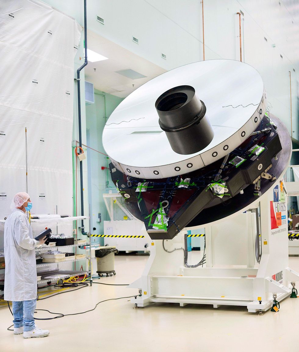 Photo of a large shiny disc on a white solid base. The structure is about twice as tall as the person in a cleanroom suit standing near it. 