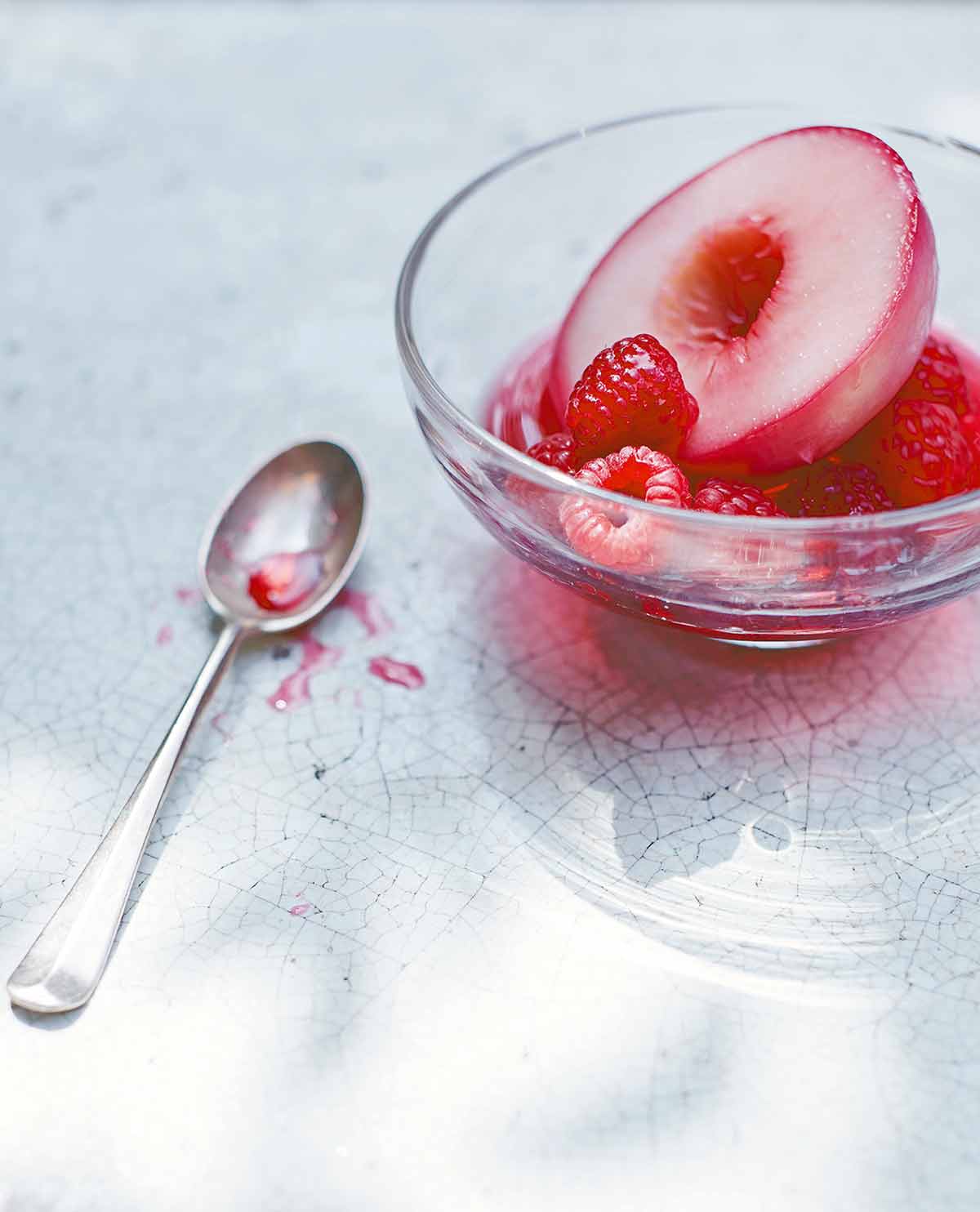 A glass bowl filled with a poached nectarine half and raspberries with a spoon on the side.