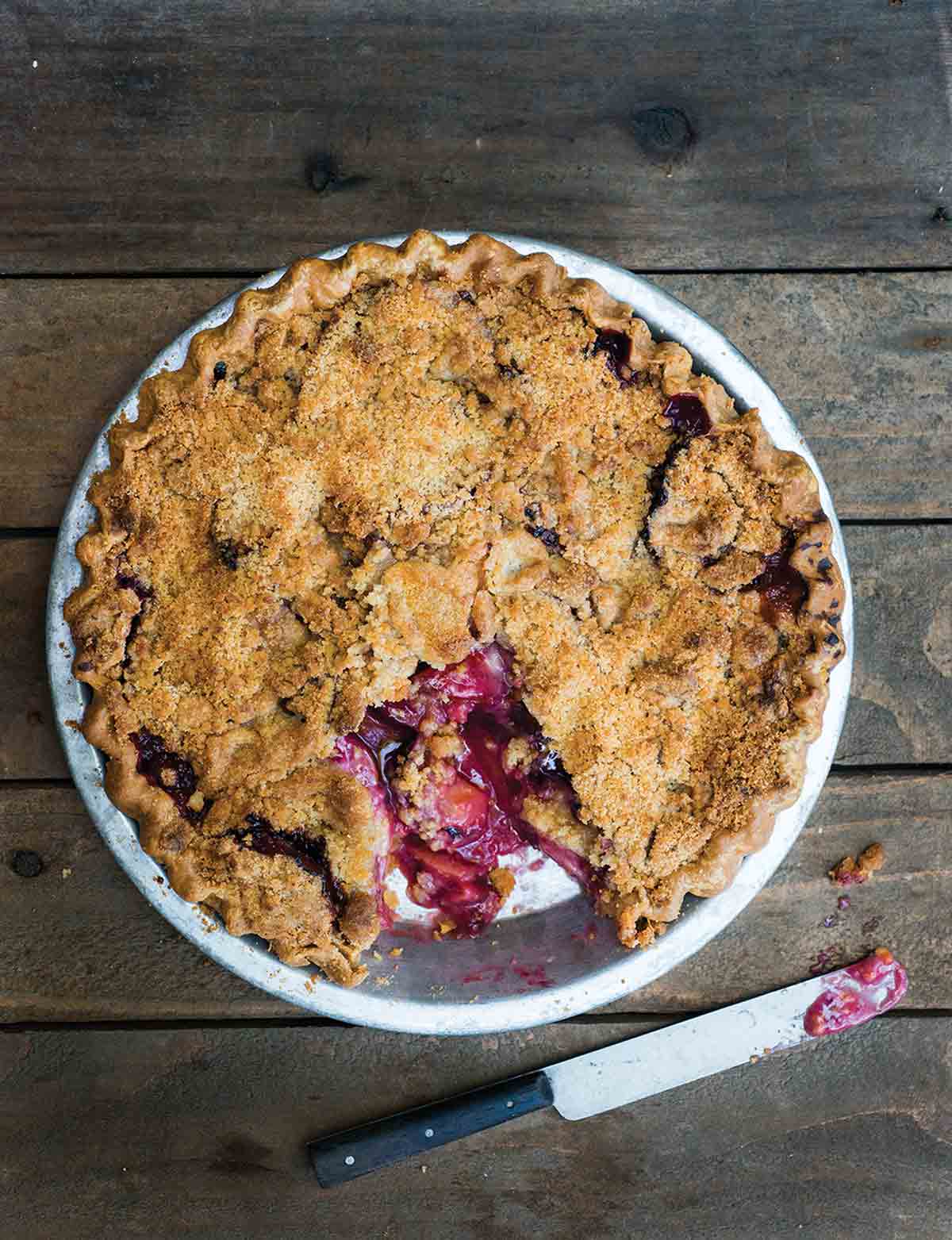 A plum pie topped with crumble with one piece missing from the pie.