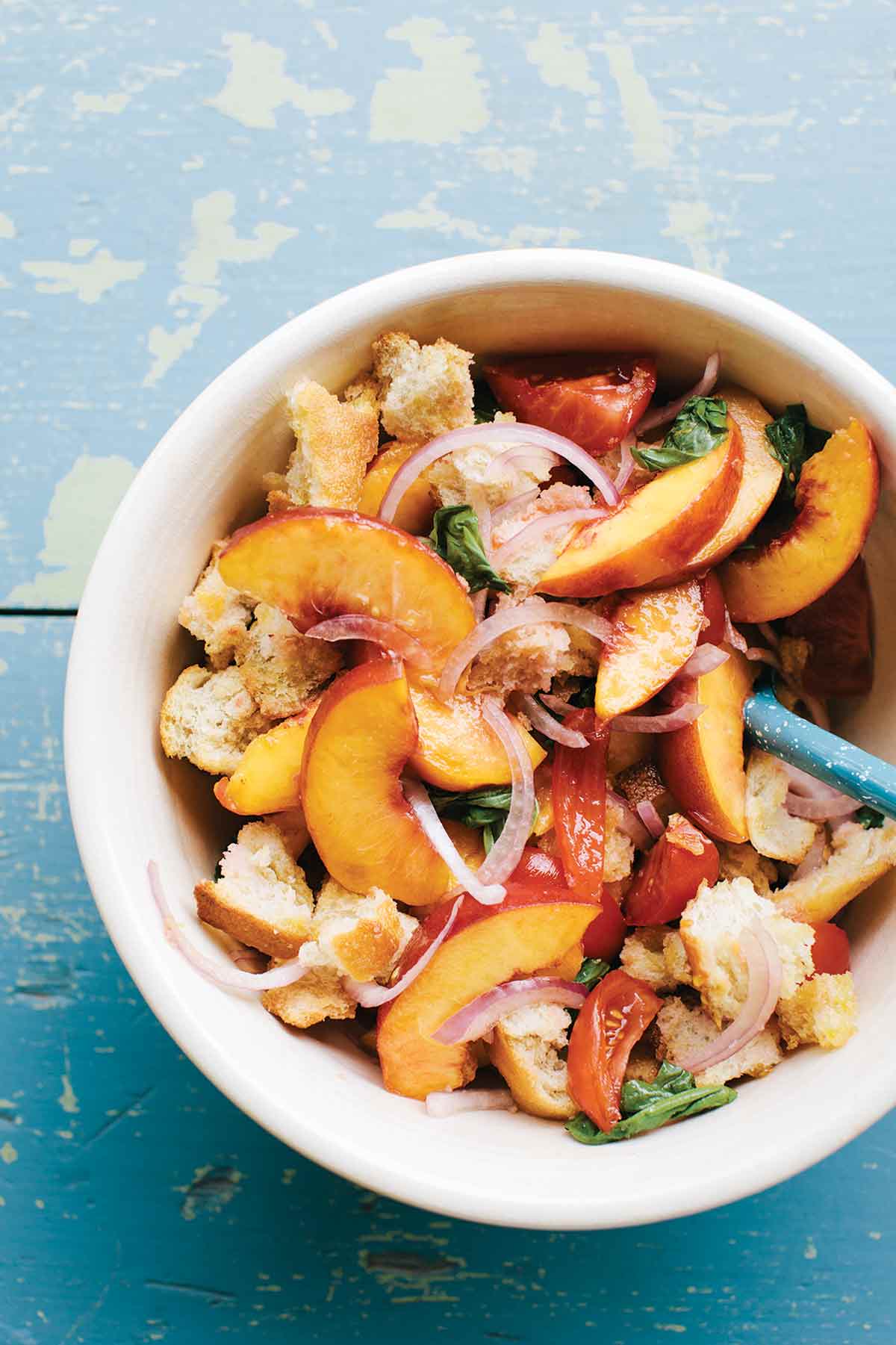 A blue background with a white bowl filled with sliced peaches, tomatoes, red onions, basil, and chunks of bread.