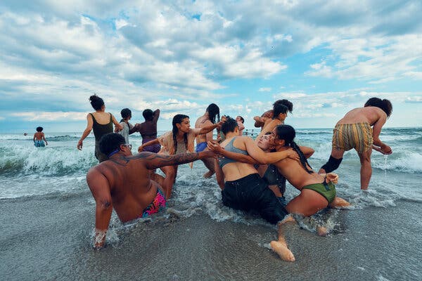 A cluster of dancers in bathing suits are at the edge of the ocean. Some hug; others link arms. A dramatically cloudy sky is overhead.