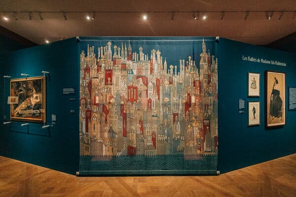 We see portions of a museum exhibition; in the center is a panel showing a painted fairy-tale city with Russian elements.