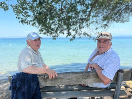 Two older men in caps sit in the shade under a tree next to the sea