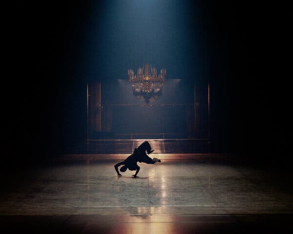 In a dimly lit room with a chandelier in the background, a silhouetted figure does a breaking move, his hands on the ground, legs out behind him.