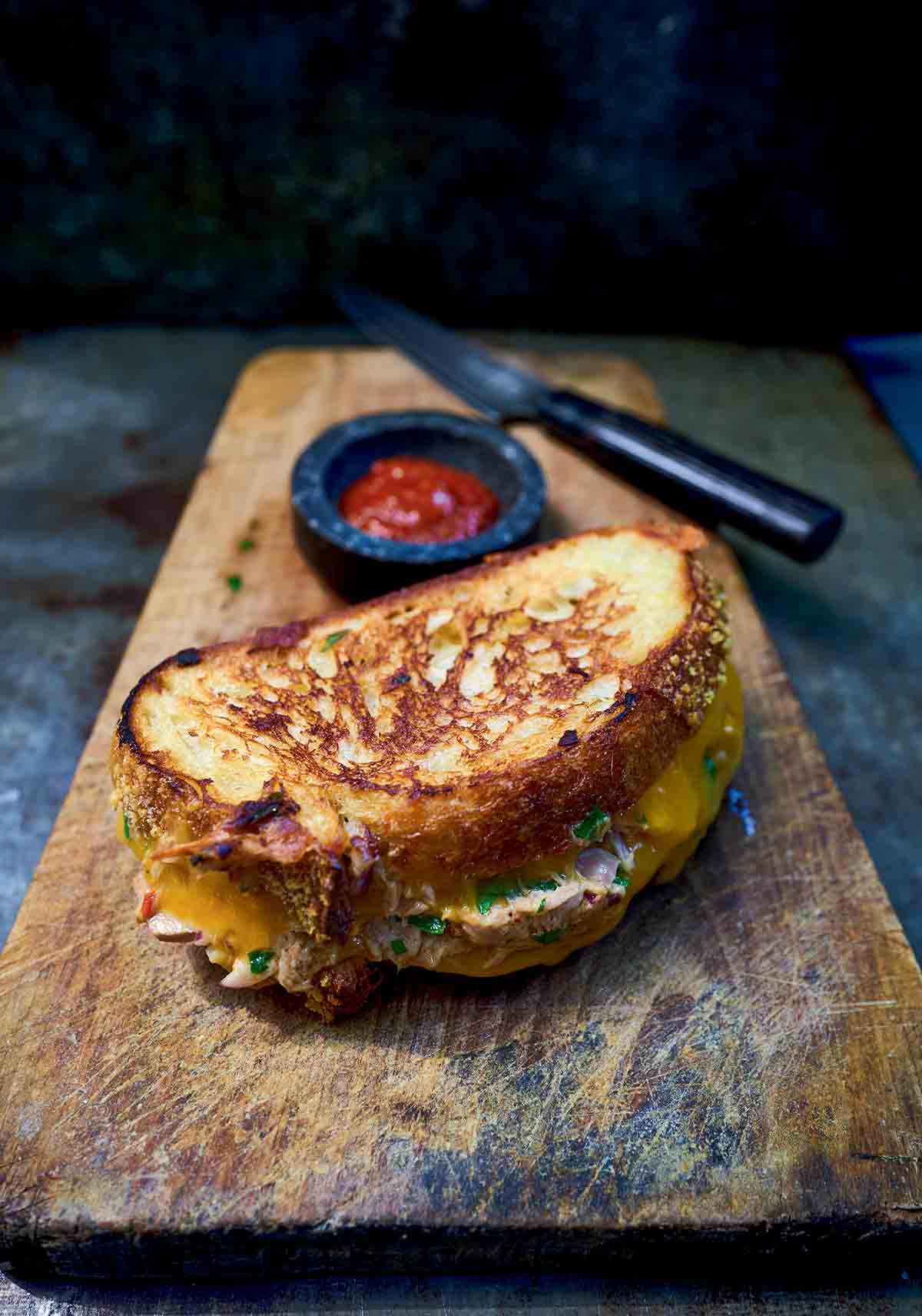 A toasted classic tuna melt on a wooden board with a dish of ketchup next to it.