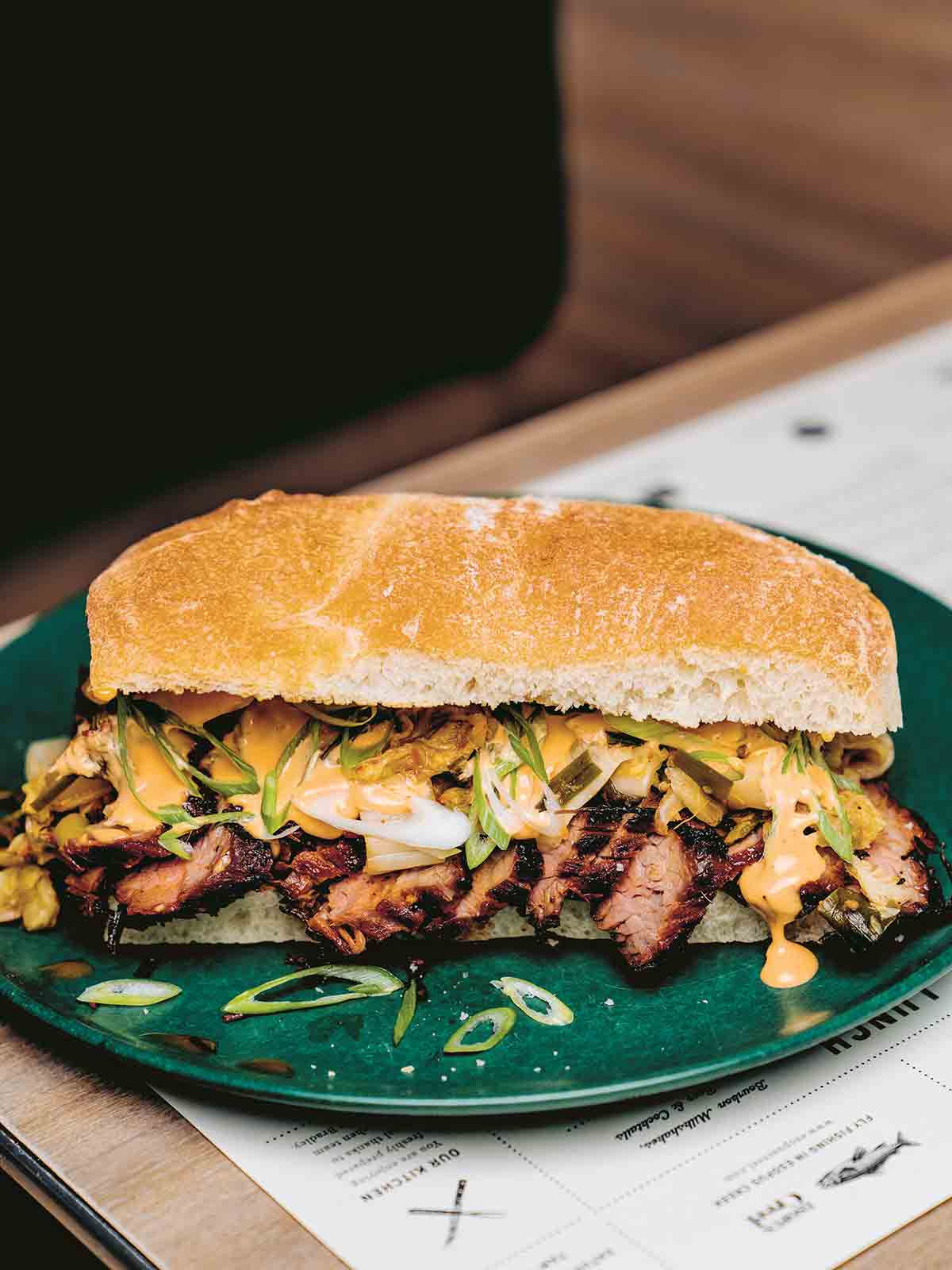 A kimchi bulgogi sandwich with sliced steak, green onions, and mayo on a roll on a green plate on top of a menu.