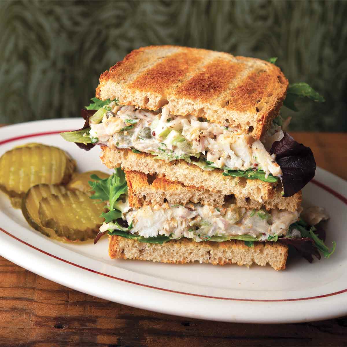 Two halves of a tuna salad sandwich stacked on top of each other with some sliced pickles on the side on a white oval plate.