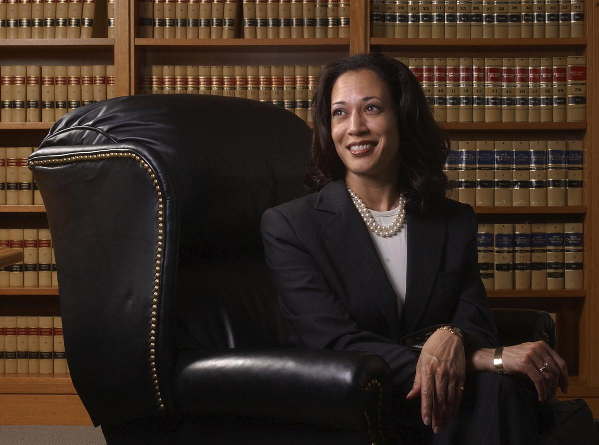 A woman in a black suit, white shirt and pearls sits in an oversized black leather chair with rows of books behind her.