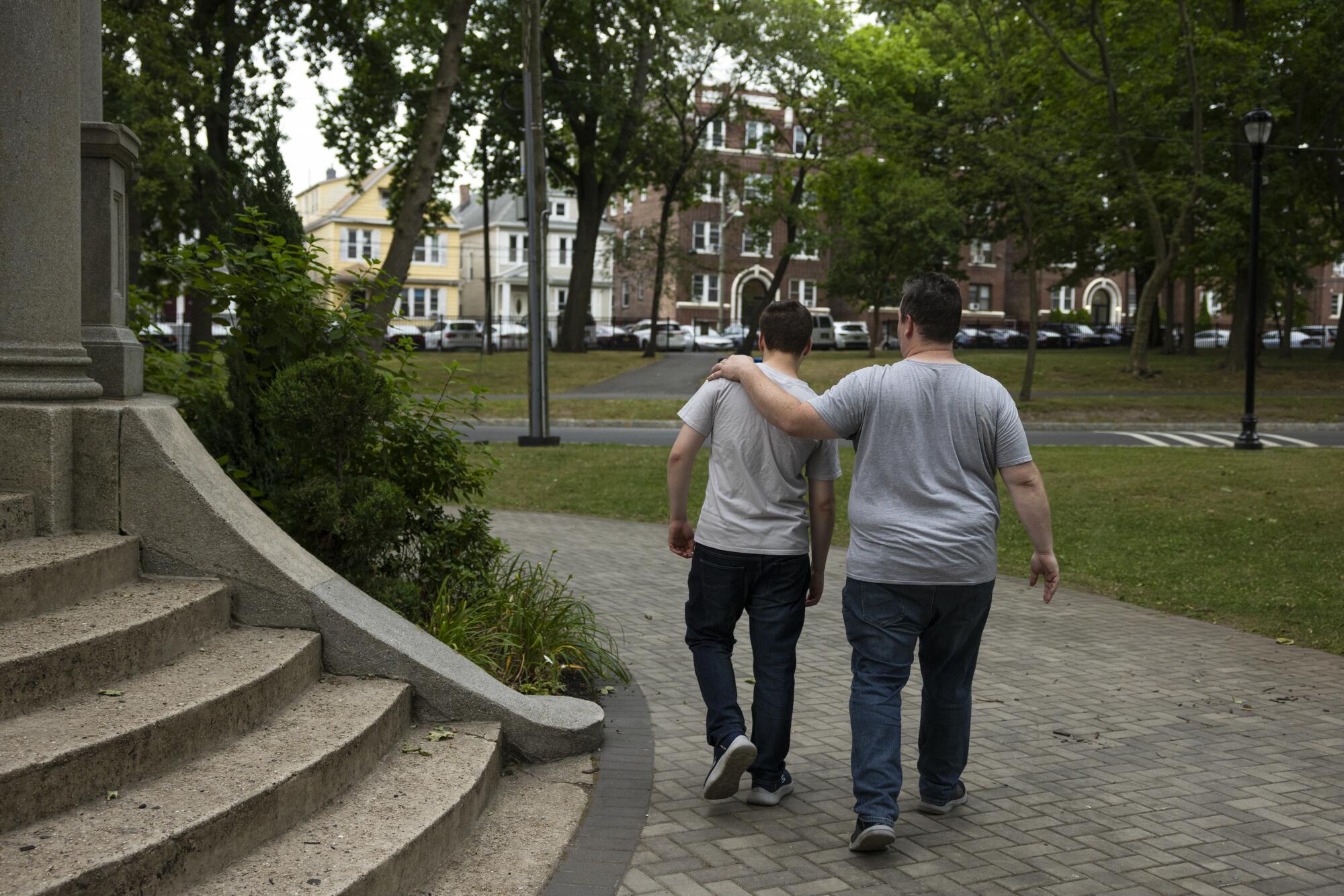 A view from behind of two men in gray T-shirts and dark pants, one with his arm around the other's shoulder, as they walk