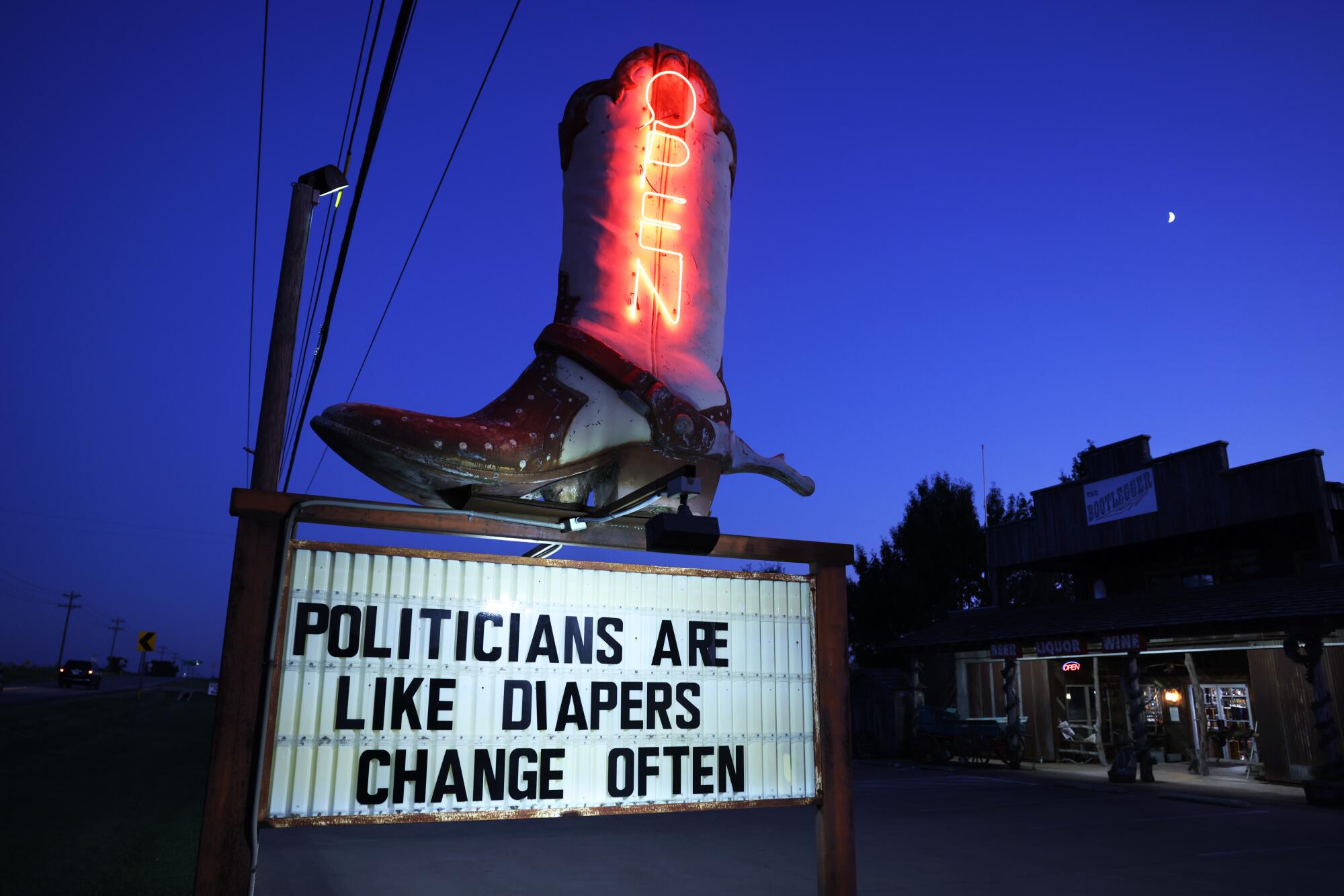 A big boot sign has neon letters reading "Open," atop a board that says, "Politicians are like diapers. Change often."  