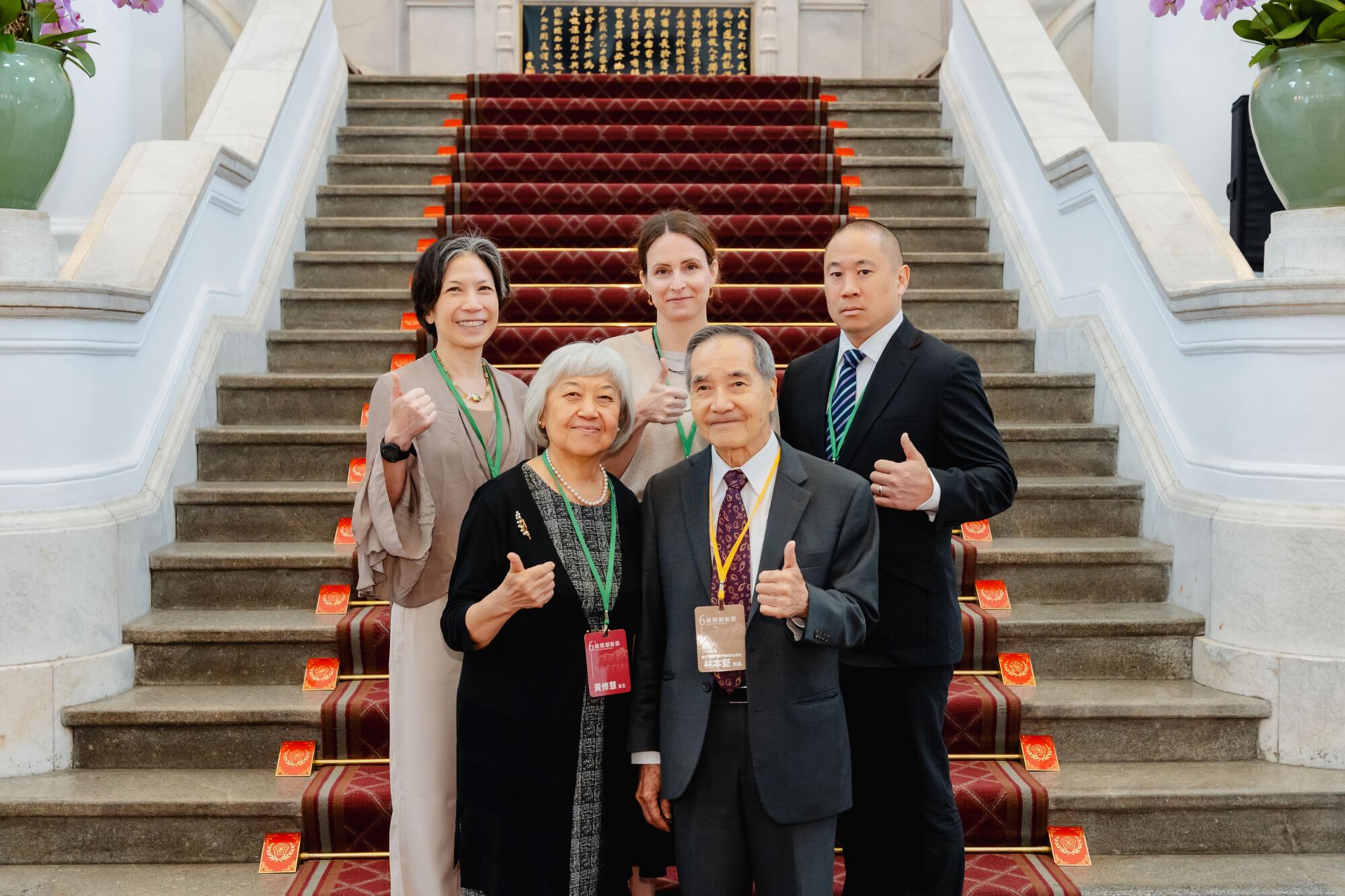 Burn Lin with several people holding up a thumb as they stand at the bottom of a carpeted staircase.