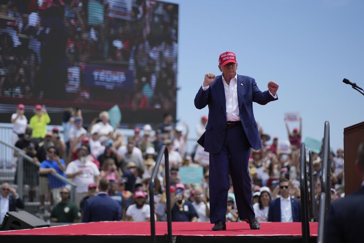 Donald Trump in a red MAGA cap, raising clenched hands while standing on an outdoor stage, a crowd in the stands behind him