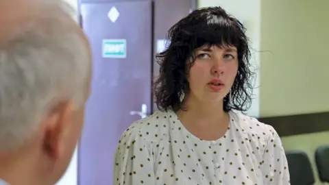 Irina, a young woman with dark curly hair and a fringe, with dangly silver earrings and wearing a white blouse with brown polka dots, looks at Steve Rosenberg, the back of whose head can be seen at the left-hand side of the picture, while speaking. 