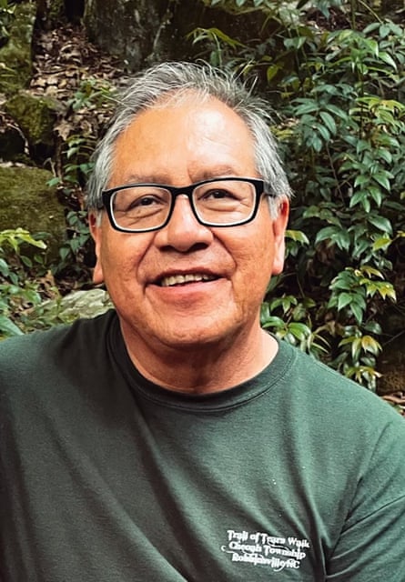 A man wearing glasses and a T-shirt standing in front of a grassy hillside smiles.