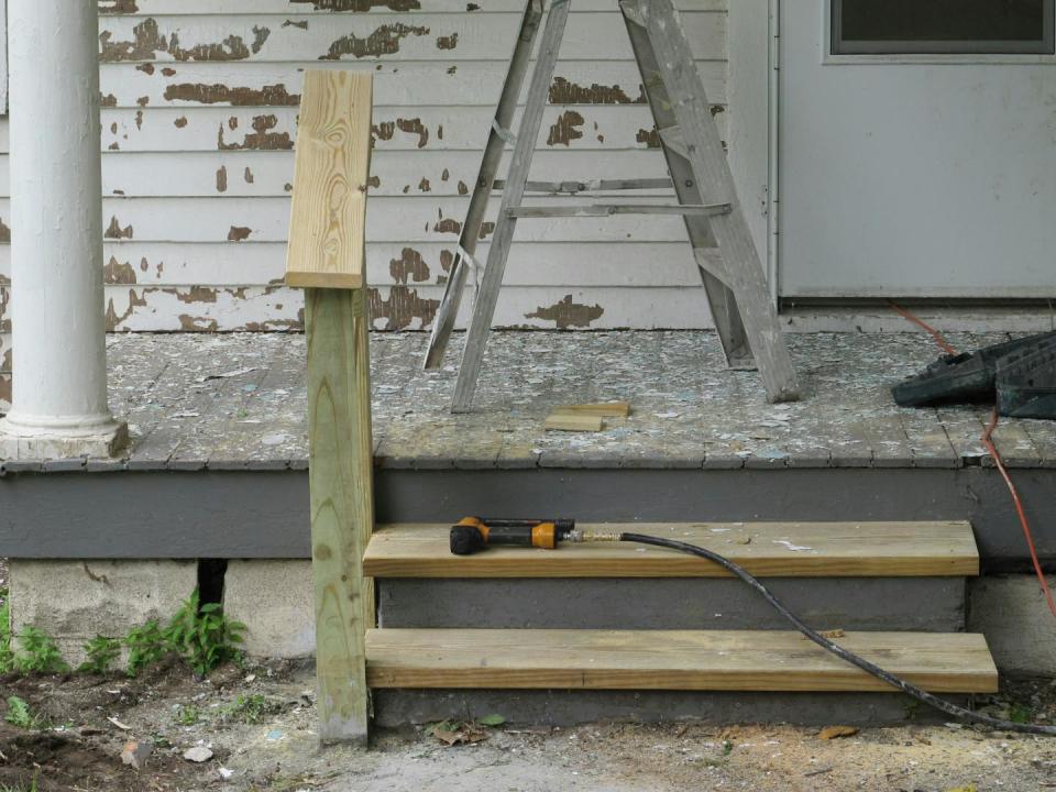 An older home with pealing paint on the exterior walls behind a porch. Construction equipment sits on a new step being built to the porch.