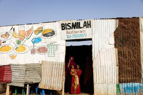 Alyona Synenko/ICRC A woman and two children are seen standing in the doorway of a cafe in Garowe refugee camp