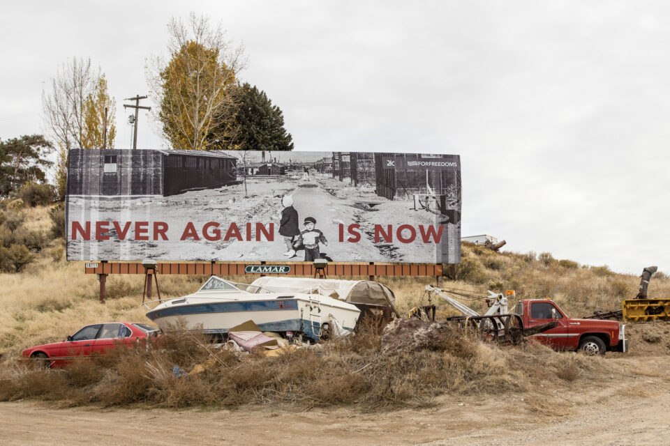 a billboard with text that reads, "NEVER AGAIN IS NOW" against a photograph of two children walking in a Japanese internment camp