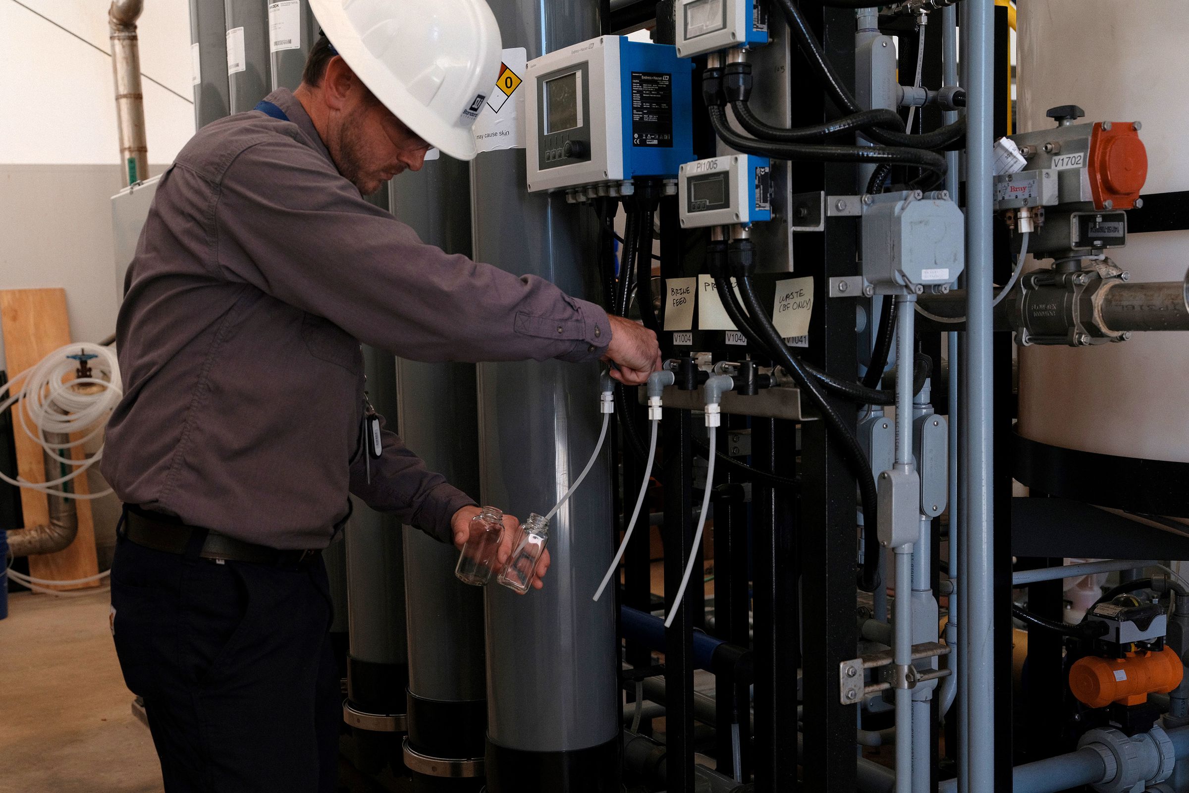 A worker wearing a hard hat inside an industrial facility turns a knob and fills a small glass bottle with liquid.