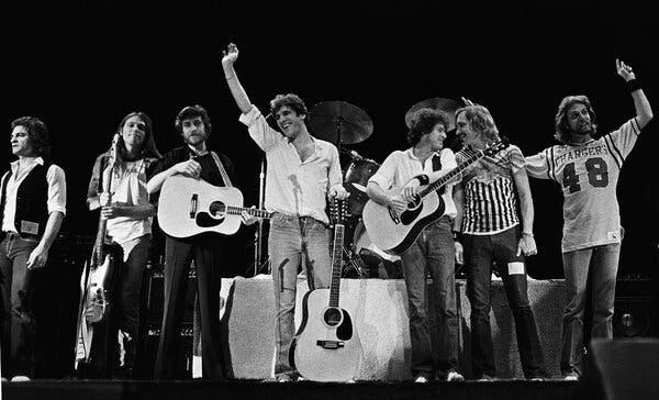 A black-and-white photo of seven rock musicians waving to the audience from the stage.