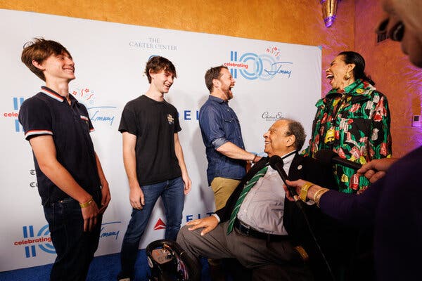 Five people laugh while positioned on a blue carpet.