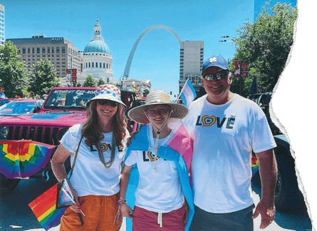 A father and mother pose with their son, as they all wear hats and rainbow shirts that say “love”, and the son has a trans flag wrapped around him