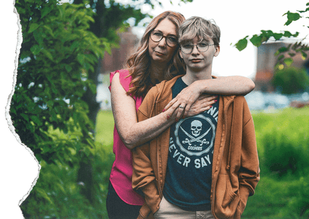 A woman embraces her son as they stand in grass next to trees