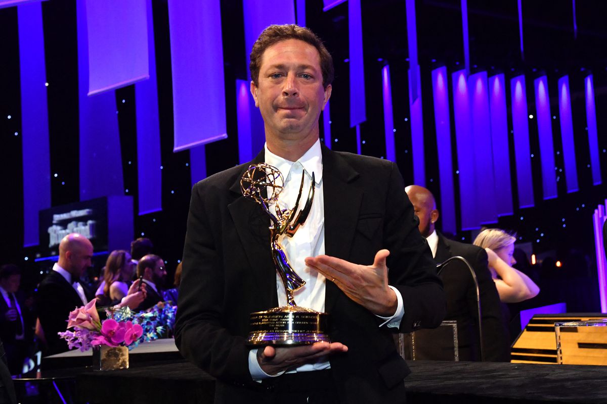 Man wearing a suit with no tie gestures at an Emmy Award in this hands inside an auditorium.