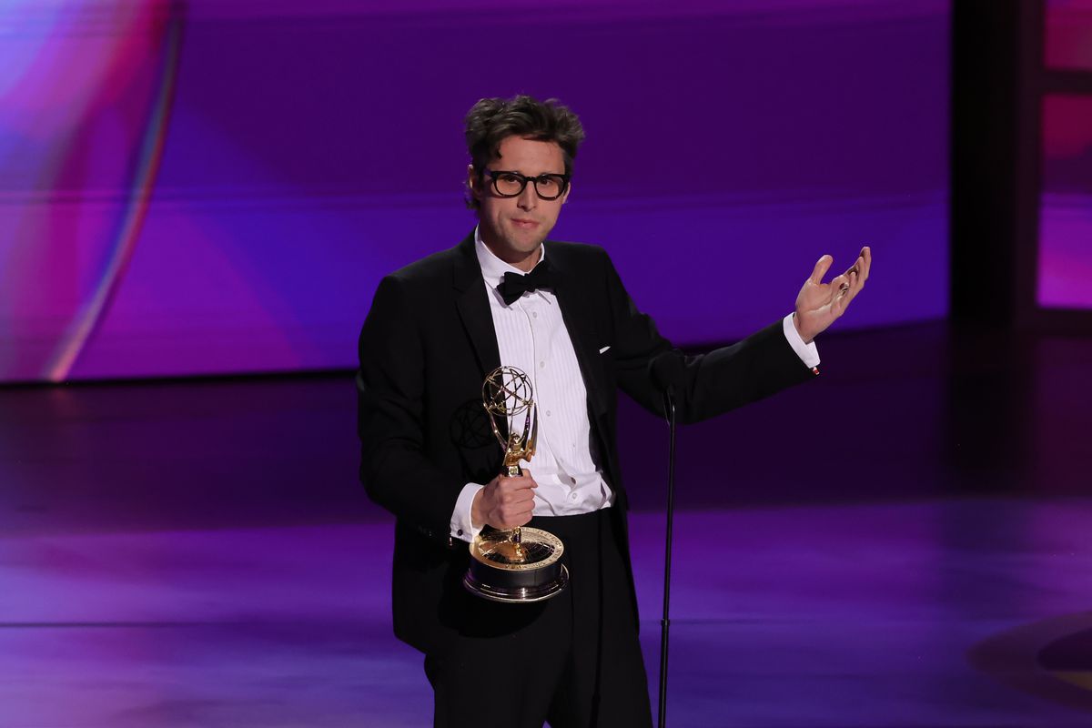 Man with glasses wearing a tux on-stage at the microphone, gesturing while holding an Emmy Award.