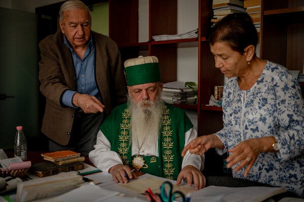 A man sitting at a desk in green and white religious clothing, including a round hat, is attended by a man and a woman. All three are viewing papers.