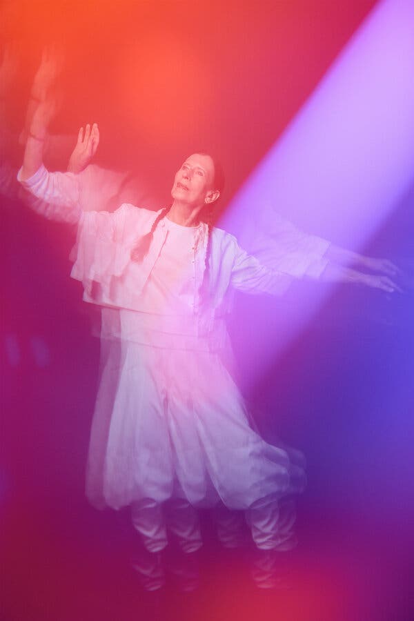 A multiple exposure portrait of Meredith Monk, dressed in white, her pigtails hanging down her front. She has her arms outstretched in a dance pose and her figure is divided by shafts of purple and red light.