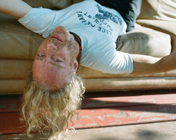 A man wearing a T-shirt hangs off the end of a couch upside-down, his long hair trailing toward the floor.