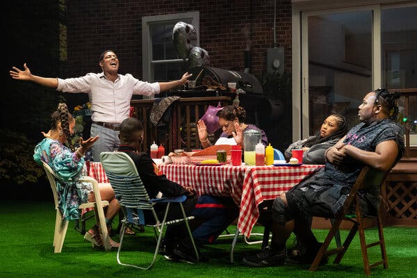 A production image show people sitting outside at a table and one man is standing with his arms outstretched.