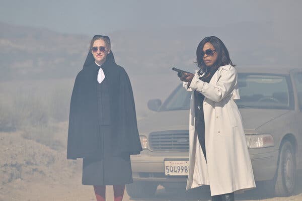 A nun and a woman in a trench coat holding a rifle, both wearing sunglasses, stand in front of a car on a dusty desert road. 