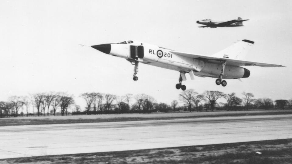  A jet plane landing on a runway accompanied by another plane behind it, in mid-air. the image is in black and white due to age. bare trees are visible in the background. 