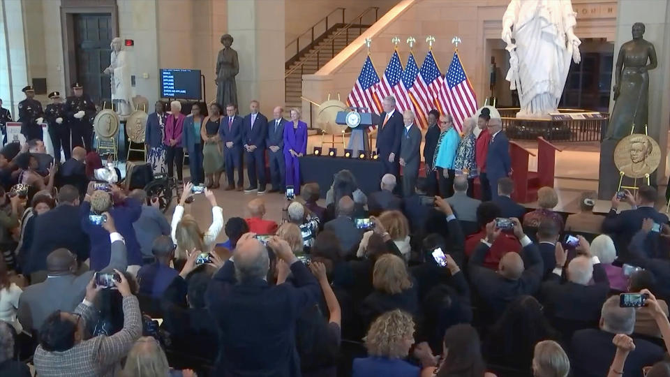  A dozen well-dressed people stand on a stage, with five american flags in the background. 