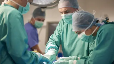 Getty Images Three surgeons in masks and green gowns operate on a patient who is lying down, unseen