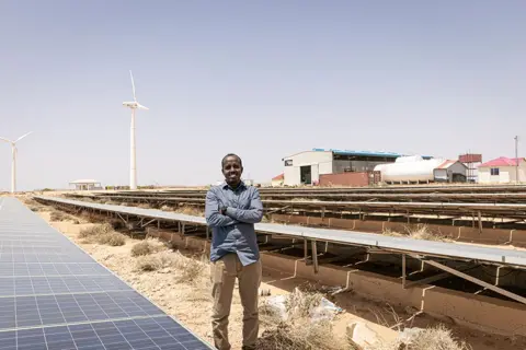 Alyona Synenko/ICRC One of the largest electricity and energy providers in Somalia, NECSOM is a  hybrid fossil fuel and green energy producer. Here a smiling man is seen standing in front of banks of solar panels, arms folded across his chest.